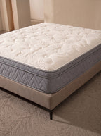 A high-profile mattress with a white quilted patterned top and gray side panels, resting on a beige upholstered bed frame in a neutral-toned room.