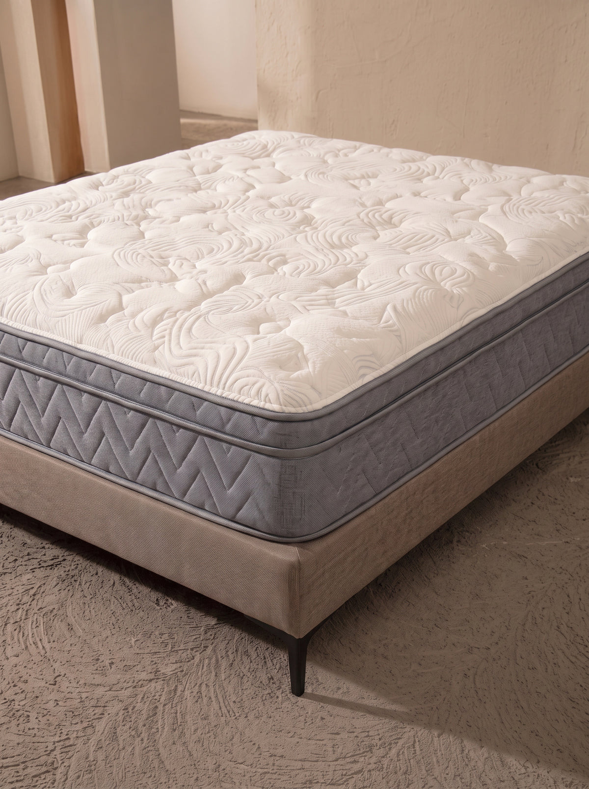 A high-profile mattress with a white quilted patterned top and gray side panels, resting on a beige upholstered bed frame in a neutral-toned room.